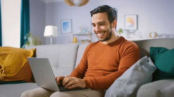 Vidám fiatalember otthon ül egy kanapén tartja Laptop az ő Lap, Böngészések az interneten, a szociális hálózatok, Vajon e-Shopping. — Stock Fotó