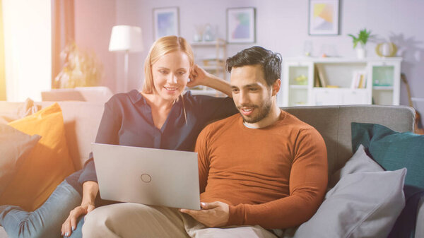 Счастливая влюбленная пара сидит дома на софе, проводит время с бойфрендом, просматривает Интернет, делает покупки в Интернете, пользуется стриминговыми сервисами. Счастливая семья в уютной гостиной. Шот с теплым солнечным светом.