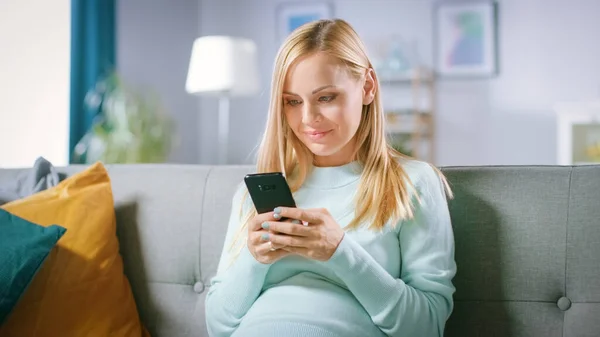 美丽快乐的孕妇坐在沙发上，用智能手机上网浏览。未来的妈妈从家里用智能手机上网购物吗？. — 图库照片