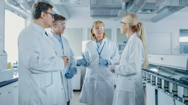 Команда вчених-дослідників має зустріч, вони обговорюють, стоячи в центрі лабораторії. Люди в інноваційній сучасній лабораторії займаються генетикою та фармацевтичними дослідженнями. — стокове фото