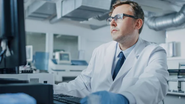 Αρσενικός ερευνητής κάθεται στο χώρο εργασίας του στο εργαστήριο, Χρησιμοποιεί τον προσωπικό υπολογιστή. Ι το ιστορικό της γενετικής, Φαρμακευτικό Κέντρο Ερευνών με καινοτόμο εξοπλισμό. — Φωτογραφία Αρχείου