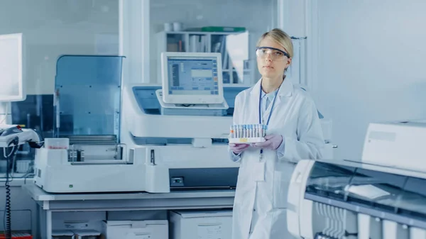 Forscherin geht mit Tablett voller Reagenzgläser voller Proben durch Labor Im Hintergrund arbeiten Menschen im Labor mit innovativen Geräten. — Stockfoto