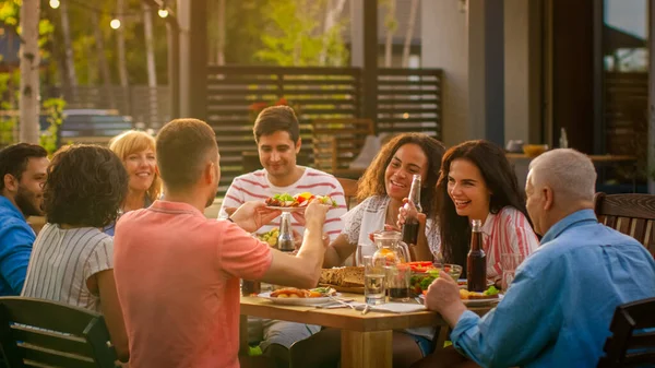 Grande festa in giardino per famiglie, riuniti a tavola parenti e amici, giovani e anziani mangiano, bevono, passano i piatti, scherzano e si divertono. — Foto Stock
