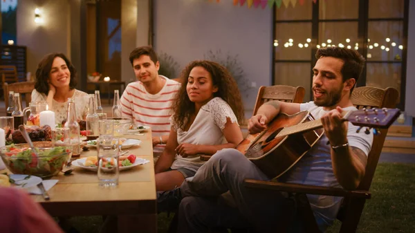 Siedząc przy stole Przystojny młody człowiek gra na gitarze dla przyjaciół. Grupa młodych ludzi słuchających muzyki na Letniej Wieczornej Imprezie Ogrodniczej. — Zdjęcie stockowe