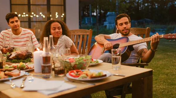 Zittend aan de eettafel Knappe jongeman speelt gitaar voor een vriend. Groep van jonge mensen die naar muziek luisteren op de zomeravond tuinfeest. — Stockfoto