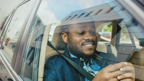 Snygg svart man reser i en bil, sitter på en passagerarstol Använder smartphone med en hand, typer Meddelande, bläddrar genom Internet. Kamera Skjuten från utanför fordonet. — Stockfoto