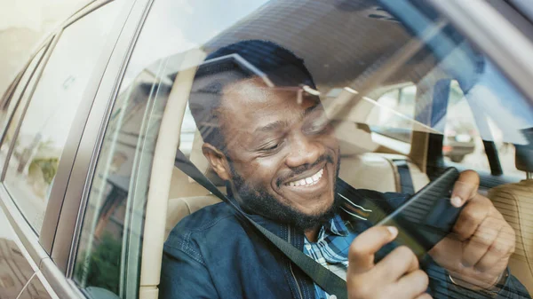 Schöner Schwarzer, der auf dem Beifahrersitz eines fahrenden Autos sitzt, spielt in einem Spiel auf einem Smartphone im Landschaftsmodus. Kamerafahrt von außerhalb des Fahrzeugs. — Stockfoto