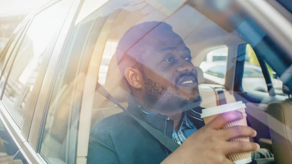 Knappe zwarte man rijdt op een passagiersstoel van een auto, kijkt in wonder uit het raam en houdt koffie. Big City View weerspiegeld in het raam. Camera gemonteerd buiten de auto. — Stockfoto