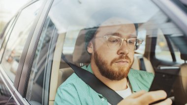 Şık bir genç adam arabaya biniyor, yolcu koltuğunda oturuyor, akıllı telefon kullanıyor, mesaj yazıyor, internete göz atıyor. Aracın dışından kamera görüntüsü.