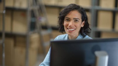 Kadın envanter müdürü masasında oturuyor ve kameraya gülümsüyor. Arkaplan Deposunda raflar karton kutu paketleriyle dolu nakliyeye hazır.