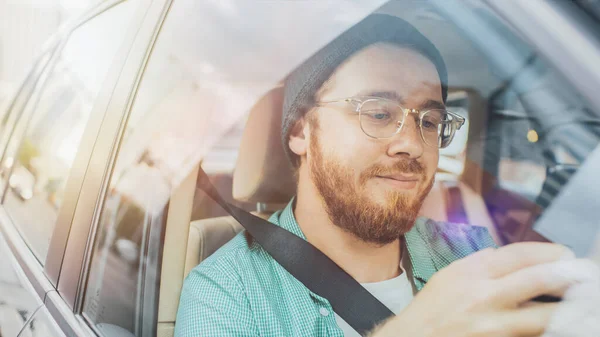 Stilvoller junger Mann, der im Auto sitzt, auf dem Beifahrersitz sitzt, Smartphone benutzt, Nachrichten tippt, im Internet surft. Kamerafahrt von außerhalb des Fahrzeugs. — Stockfoto