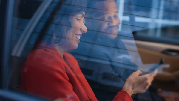 Die schöne Frau und ihr männlicher Partner sitzen auf dem Rücksitz eines Autos, teilen sich den Smartphone-Bildschirm und unterhalten sich und haben Spaß. Sein Abend in der Großstadt. — Stockfoto