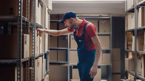 El trabajador guapo del almacén está buscando una caja de cartón en un estante con otros paquetes. — Foto de Stock