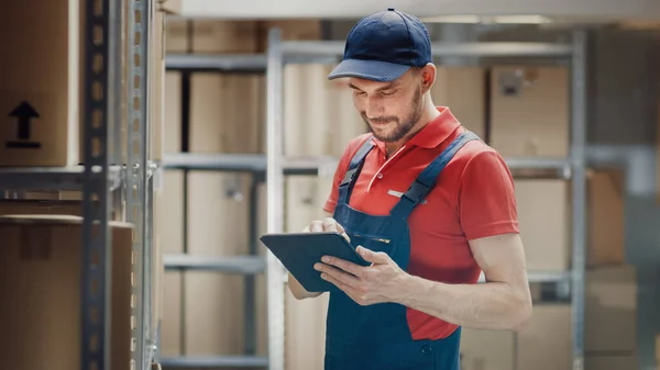 El trabajador guapo del almacén utiliza la tableta digital para comprobar el stock, en las cajas de cartón de pie de los estantes. — Foto de Stock