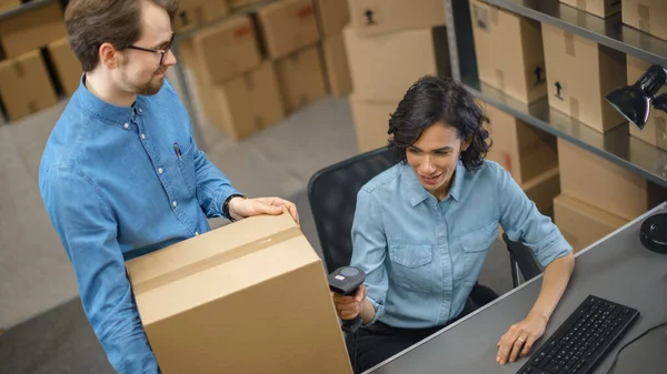 El gestor de inventario femenino escanea la caja de cartón y con el escáner de código de barras, el trabajador masculino sostiene el paquete. En las filas de fondo de cajas de cartón con productos listos para el envío. — Foto de Stock