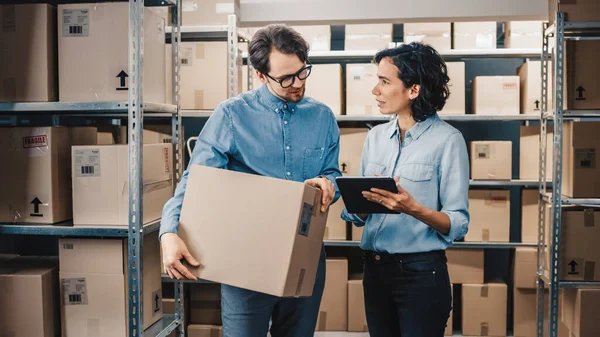 Female Inventory Manager Shows Digital Tablet Information to a Worker Holding Cardboard Box, They Talk and Do Work. In the Background Stock of Parcels with Products Ready for Shipment. — Stock Photo, Image
