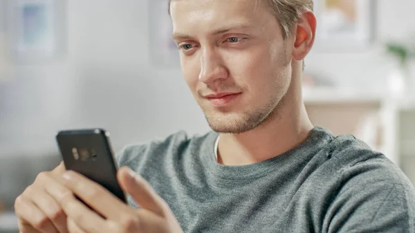 Portrett av den kjekke unge mannen som bruker smarttelefon, nettlesing, sjekker sosiale nettverk mens han sitter hjemme. – stockfoto