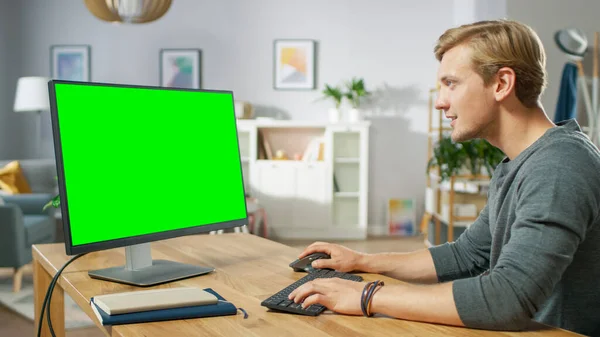 Bonito jovem trabalha em um computador pessoal verde Mock-up tela enquanto sentado em sua mesa no aconchegante apartamento. — Fotografia de Stock