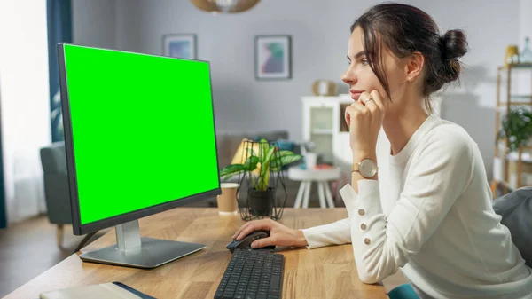 Menina bonita trabalha em um computador pessoal verde da tela Mock-up enquanto sentado em sua mesa no aconchegante apartamento. — Fotografia de Stock