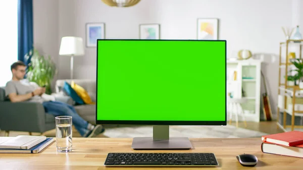 Modern Personal Computer with Green Mock-up Screen Display Standing on the Desk in the Cozy Living Room. A Man with Mobile Phone Relaxes in His Chair. — Stock Photo, Image