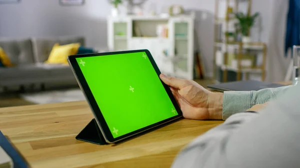 Over the Shoulder Shot of a Man Holding and Watching Green Mock-up Screen Digital Tablet Computer While Sitting at the Desk. No fundo aconchegante sala de estar. — Fotografia de Stock