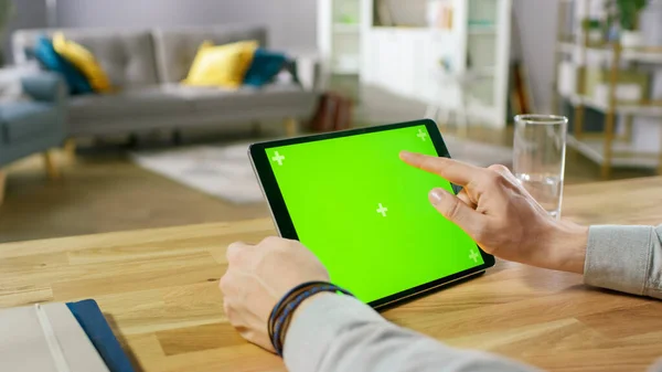 Man Holding and Using Hand Gestures on Green Mock-up Screen Digital Tablet Computer While Sitting at the Desk. Homem a comprar coisas ou a navegar pela Internet. No fundo aconchegante sala de estar. — Fotografia de Stock