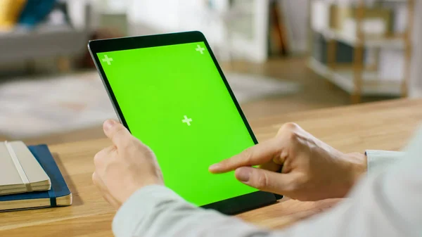 Uomo che utilizza gesti mano su Green Mock-up schermo computer tablet digitale in modalità ritratto mentre seduto alla sua scrivania. Sullo sfondo accogliente soggiorno. — Foto Stock