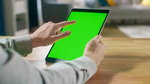 Uomo che utilizza gesti mano su Green Mock-up schermo computer tablet digitale in modalità ritratto mentre seduto alla sua scrivania. Sullo sfondo accogliente soggiorno. — Foto Stock