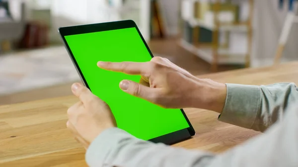 Man Using Hand Gestures on Green Mock-up Screen Digital Tablet Computer in Portrait Mode miközben ül az asztalánál. A háttérben Hangulatos nappali. — Stock Fotó