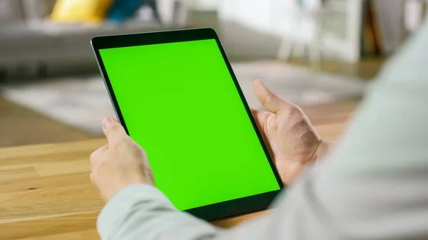 Man Using Green Mock-up Screen Digital Tablet Computer in Portrait Mode while Seitting at His Desk. V pozadí útulný obývací pokoj. — Stock fotografie