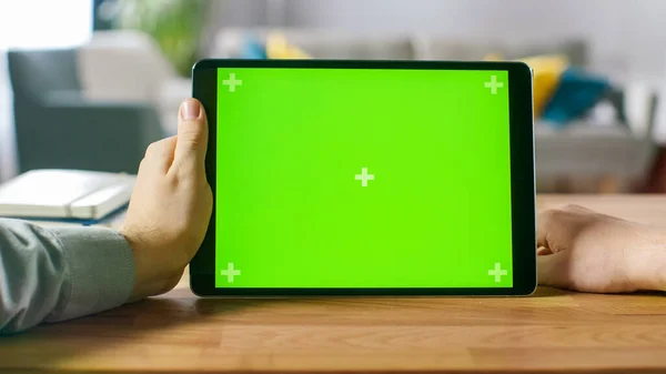 Ego-Shot eines Mannes mit grünem Mock-up Screen Digital Tablet Computer im Landscape-Modus, während er an seinem Schreibtisch sitzt. Im Hintergrund gemütliches Wohnzimmer. — Stockfoto