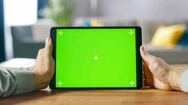 Nahaufnahme eines Mannes mit grünem Mock-up Screen Digital Tablet Computer im Landscape-Modus, während er an seinem Schreibtisch sitzt. Im Hintergrund gemütliches Wohnzimmer. — Stockfoto