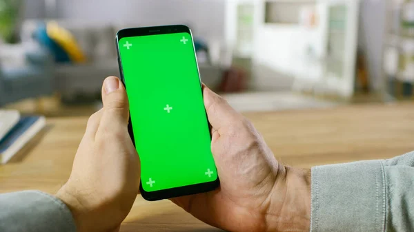 Közelkép egy férfiról, aki kezében tartja a Modern Zöld Mock-up Screen Smartphone-t. Modern high-end eszközt használok. A háttérben Hangulatos nappali vagy Home Office. — Stock Fotó
