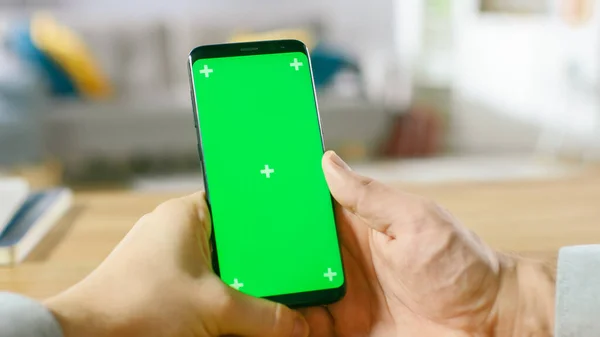 Gros plan d'un homme tenant un téléphone intelligent vert maquillé dans ses mains. Utiliser un appareil sans fil pour naviguer sur Internet. En arrière-plan Salon confortable ou bureau à domicile. — Photo