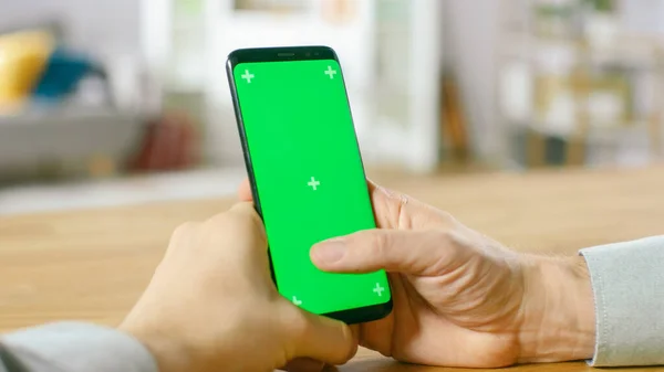 Зблизька людина тримає зелений макет-екран Смартфон у своїх руках. Використання бездротового пристрою для проходження через Інтернет. На задньому плані затишна вітальня або домашнє бюро. — стокове фото