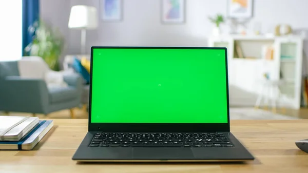 Laptop moderno com tela verde Mock-up em pé sobre a mesa na sala de estar acolhedora. Homem com telefone móvel caminha através de seu apartamento. — Fotografia de Stock