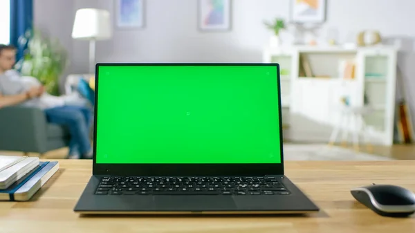 Moderner Laptop mit grünem Bildschirmmotiv, der auf dem Schreibtisch im gemütlichen Wohnzimmer steht. Mann mit Handy setzt sich zum Ausruhen auf einen Stuhl. — Stockfoto