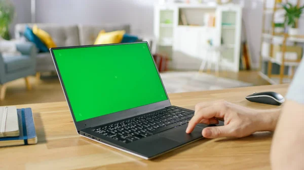 Mann benutzt Laptop mit grünem Bildschirm, während er am Schreibtisch in seinem gemütlichen Wohnzimmer sitzt. — Stockfoto