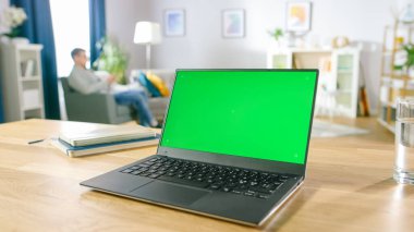 Yeşil Model Ekranlı Modern Laptop Samimi Oturma Odasındaki Masanın Üzerinde Duruyor. Cep telefonu olan adam dinlenmek için sandalyeye oturdu..