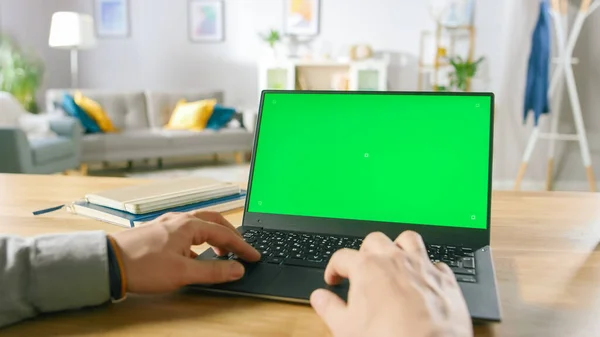 Close-up van een man maakt gebruik van laptop met groene Mock-up scherm tijdens het zitten aan de balie in zijn gezellige woonkamer. — Stockfoto
