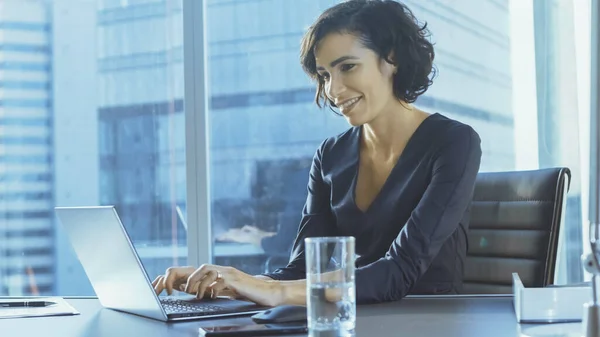 A executiva confiante trabalha em um laptop sentado em sua mesa no escritório moderno com vista para a cidade grande. Sorrindo bem sucedido Busiesswoman usa laptop. — Fotografia de Stock