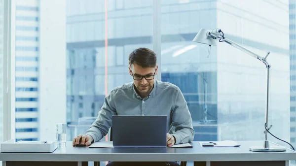 Schöner junger Geschäftsmann, der mit Laptop im Büro an seinem Schreibtisch sitzt, hebt die Hände und feiert beeindruckende Haushaltsergebnisse. Im Hintergrundfenster mit Blick auf die Stadt. — Stockfoto