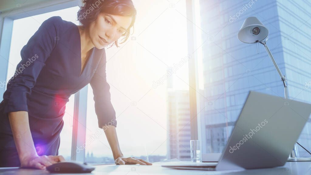 Low Angle Shot of Confident Businesswoman Leans on Her Office Desk, Looks at Documents and Using Laptop. Successful Woman Doing Business. Sun Flare Behind Her.