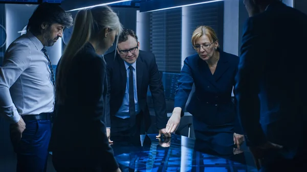 Diverse Team of Government Intelligence Agents Standing Around Digital Touch Screen Table og Tracking Suspect, Senior Officer gør Interactive Gesturing. Stort mørkt overvågningsrum. - Stock-foto