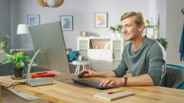 Masasında otururken Kişisel Bilgisayarda Çalışan Odaklanmış Genç Adamın Portresi. Arka plandaki rahat oturma odasında..