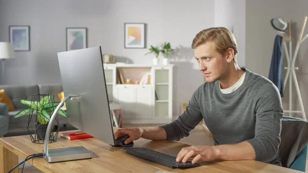 Retrato del joven enfocado trabajando en una computadora personal mientras está sentado en su escritorio. En el fondo acogedor salón. — Foto de Stock