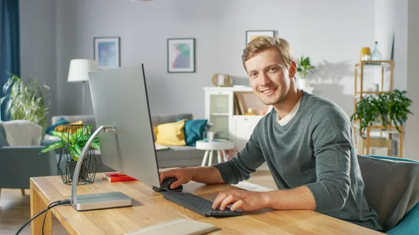 Πορτρέτο του Εστιασμένου Νέου που εργάζεται σε έναν προσωπικό υπολογιστή, ενώ κάθεται στο γραφείο του, χαμογελώντας και κοιτάζοντας την κάμερα. Στο φόντο άνετο σαλόνι. — Φωτογραφία Αρχείου