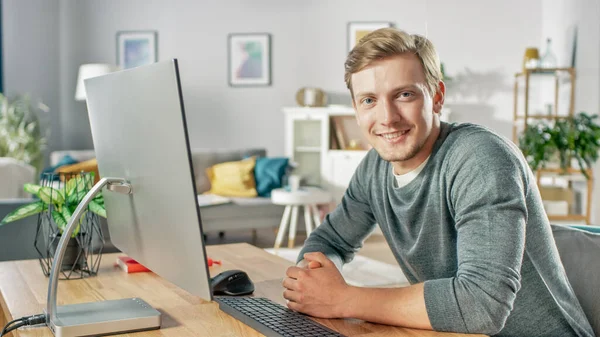 Πορτρέτο του Εστιασμένου Νέου που εργάζεται σε έναν προσωπικό υπολογιστή, ενώ κάθεται στο γραφείο του, χαμογελώντας και κοιτάζοντας την κάμερα. Στο φόντο άνετο σαλόνι. — Φωτογραφία Αρχείου
