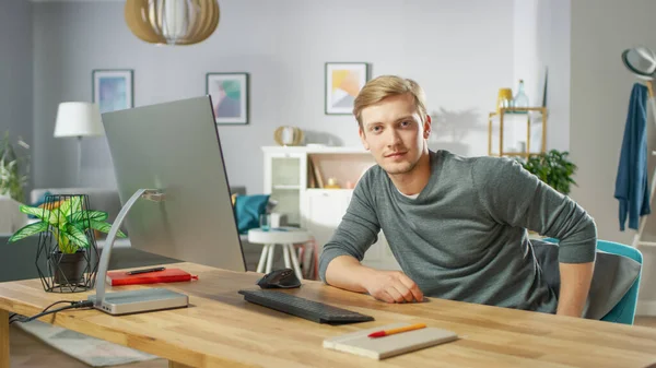 Portrait du beau jeune homme regardant la caméra assis à son bureau avec un ordinateur personnel. En arrière-plan Salon confortable. — Photo