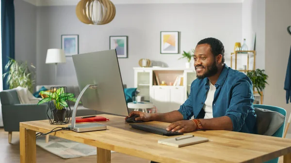 Όμορφος μαύρος άνθρωπος εργάζεται σε έναν προσωπικό υπολογιστή, ενώ κάθεται στο γραφείο του άνετο σαλόνι του. — Φωτογραφία Αρχείου